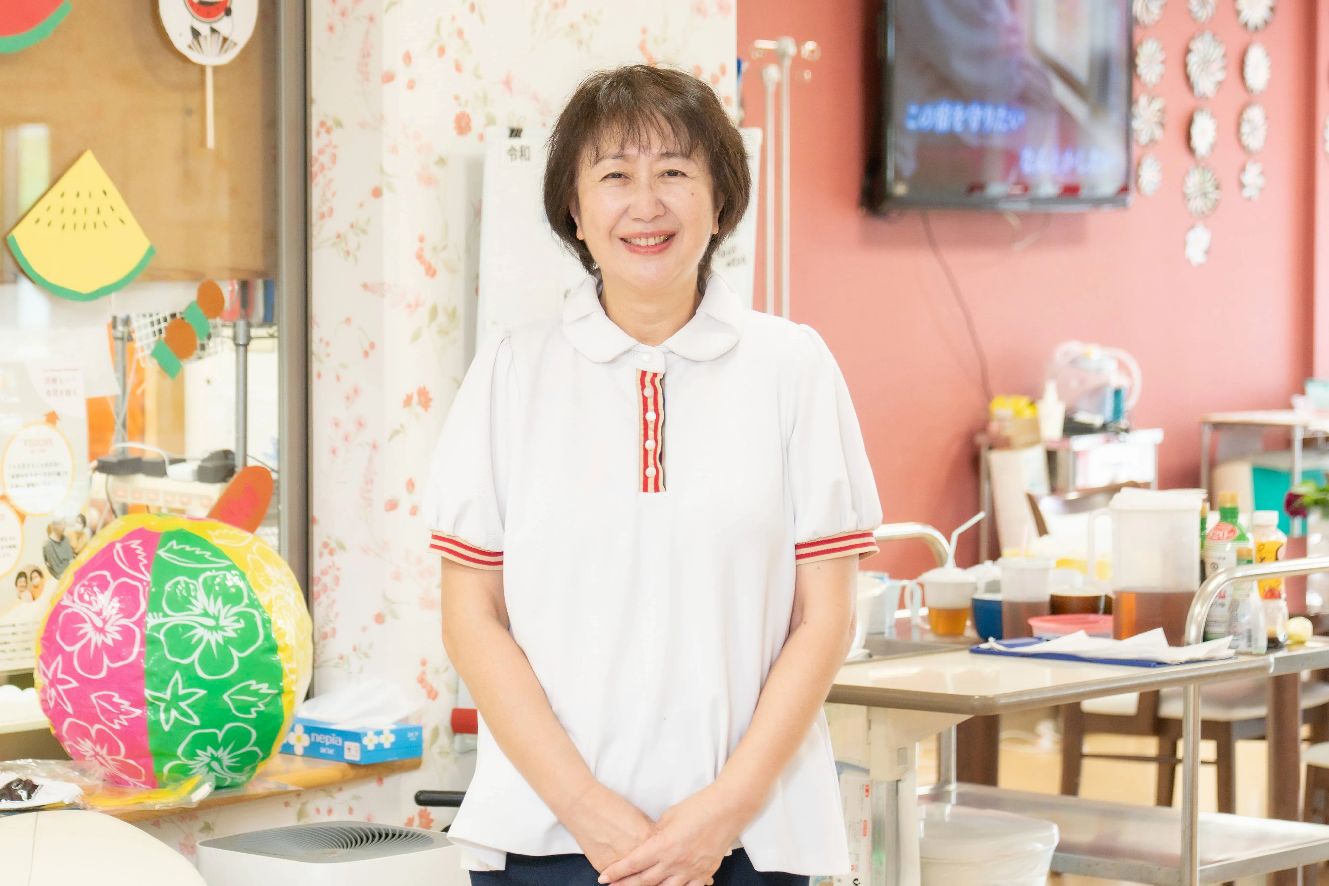介護施設のスタッフが活動室で笑顔で立つ。壁には児童向けの装飾や玩具が見える。